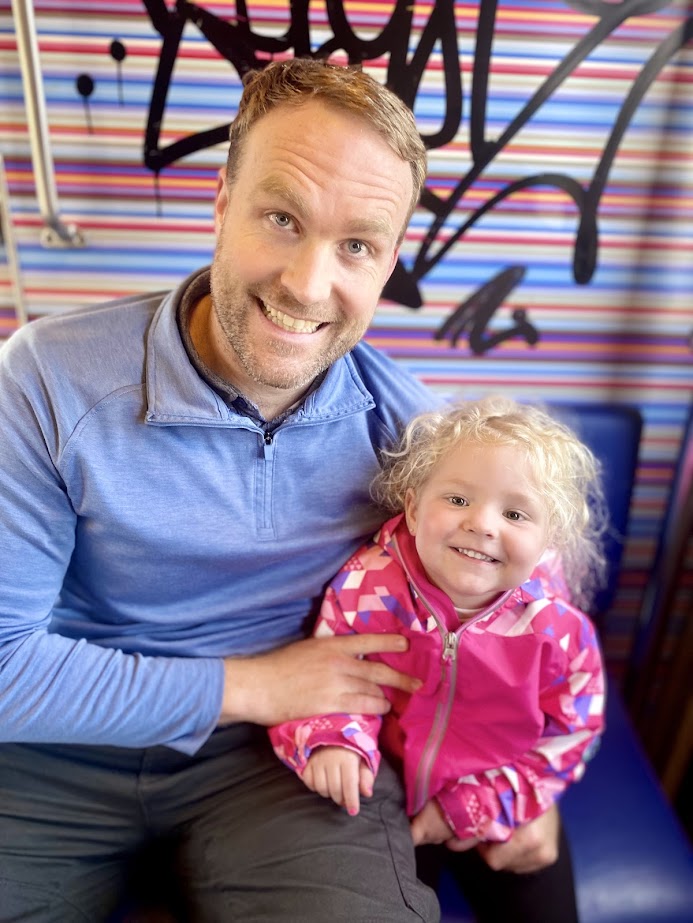 A smiling man and a young girl, both sitting on a bench in a colorful environment with stripes and graffiti art in the background.
