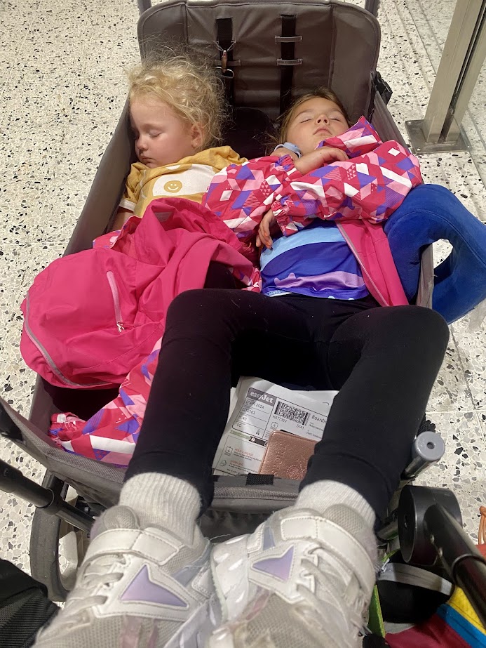 Two children sleeping in a stroller at an airport, surrounded by colorful jackets and a backpack.