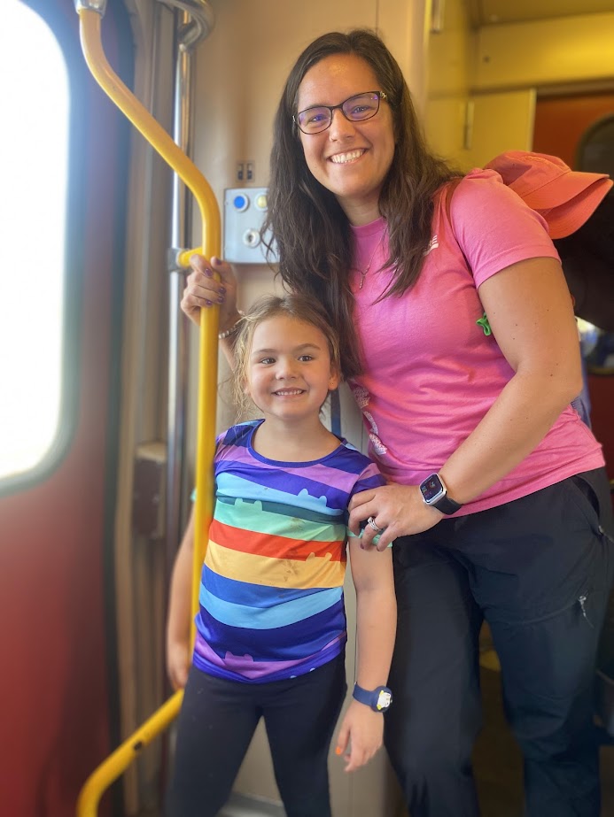 A smiling woman and a young girl are standing together in a train, holding onto a yellow pole. The woman is wearing a pink shirt, and the girl is in a colorful striped shirt.
