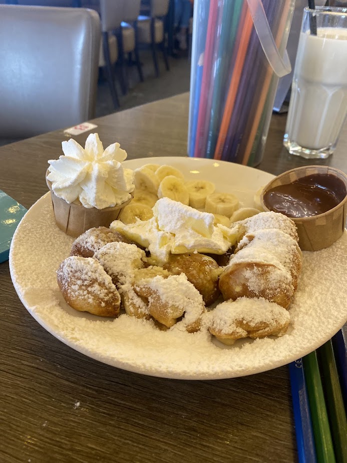 A dessert plate featuring powdered sugar-dusted dough balls, a serving of whipped cream, banana slices, and a small cup of chocolate sauce.