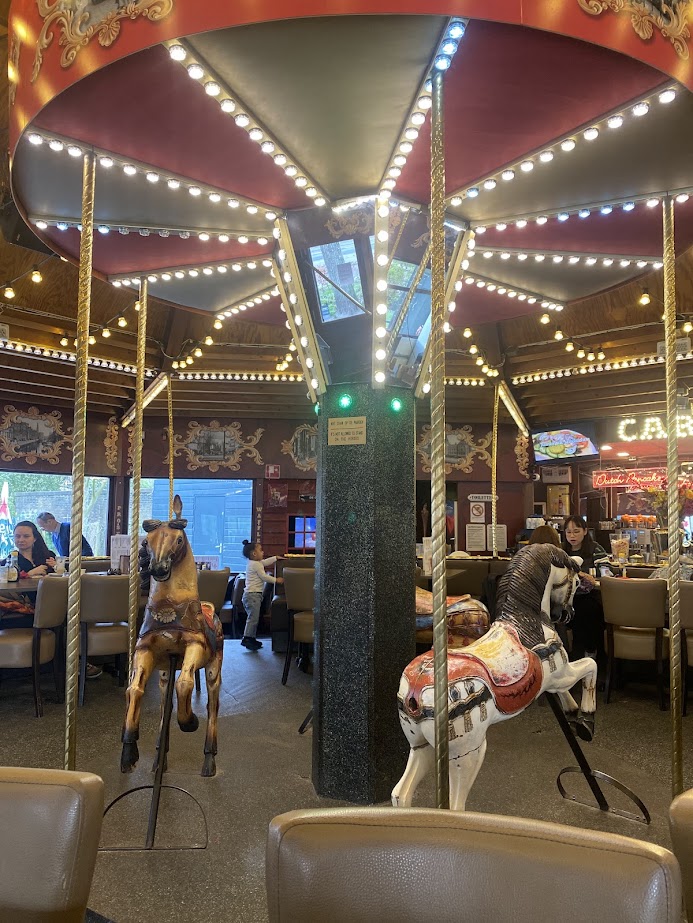Interior of a carousel-themed restaurant featuring two carved horse figures, string lights, and diners in the background.