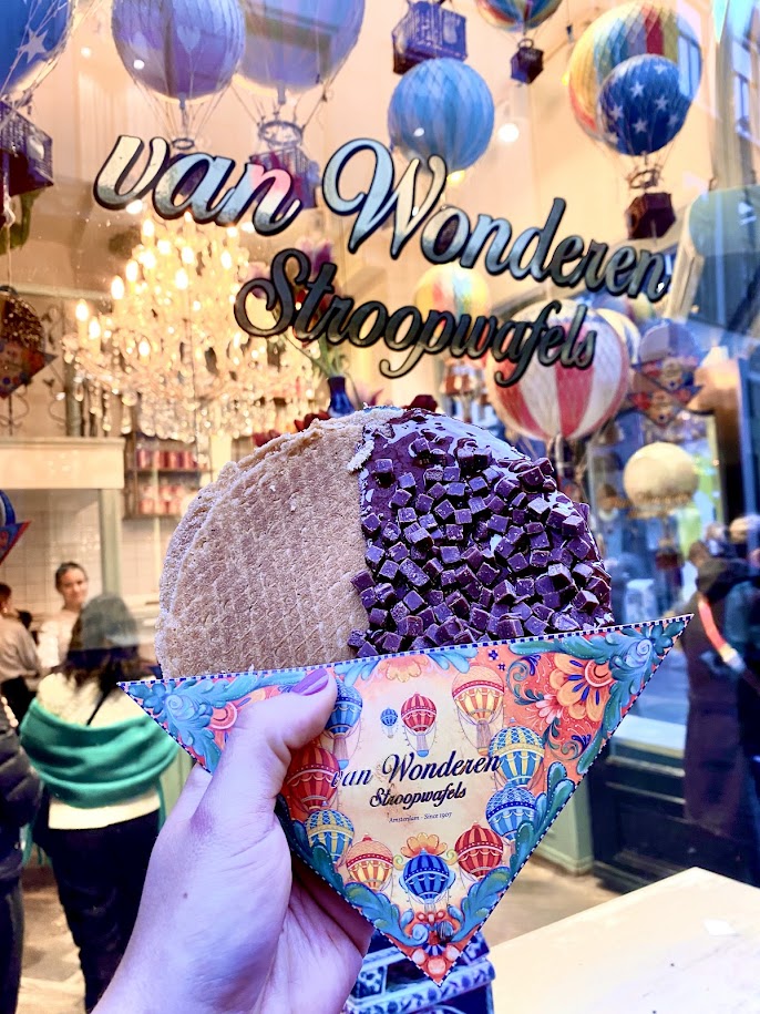 A hand holding a large stroopwafel with a chocolate and decorative topping, in front of a shop window displaying the name 'van Wonderen Stroopwafels' and colorful hot air balloons.