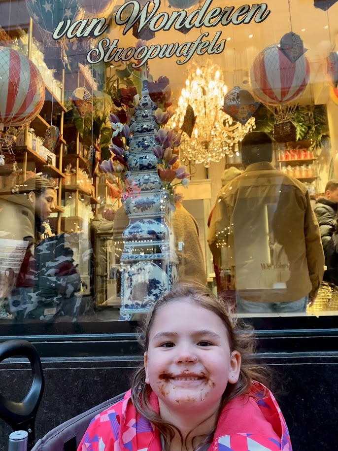 A young girl with chocolate on her face smiles in front of a shop window displaying decorative items and a chandelier. The sign above reads 'van Wonderen Stroopwafels.'