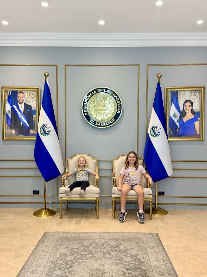 Two children sitting on chairs in a room decorated with the flags of El Salvador and presidential portraits.