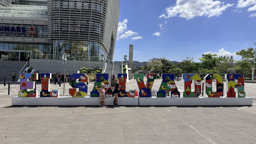 Colorful 'EL SALVADOR' sign with artistic designs in front of a modern building, two children posing beside it on a sunny day.