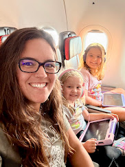 A woman and two young girls smiling for a selfie while sitting in an airplane, with the airplane window in the background.