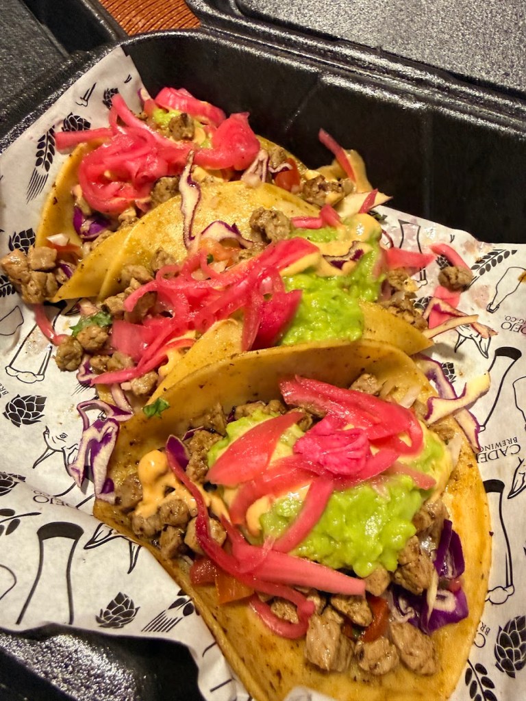 Three tacos filled with chopped meat, guacamole, pickled onions, and shredded cabbage, served in a takeout container.