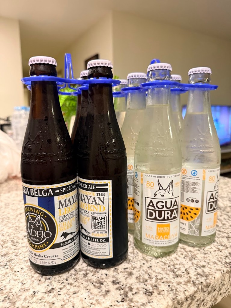 A close-up image of several bottles of Cadejo Brewing Company drinks, including spiced ale and flavored water, held together by a blue bottle carrier on a granite countertop.