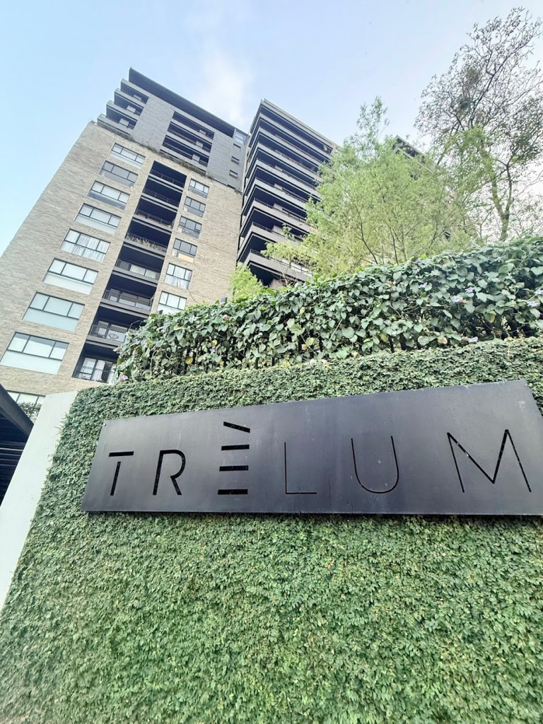 View of a modern apartment building with balconies and greenery in the foreground, featuring the name 'TRÊLUM' on a sign.