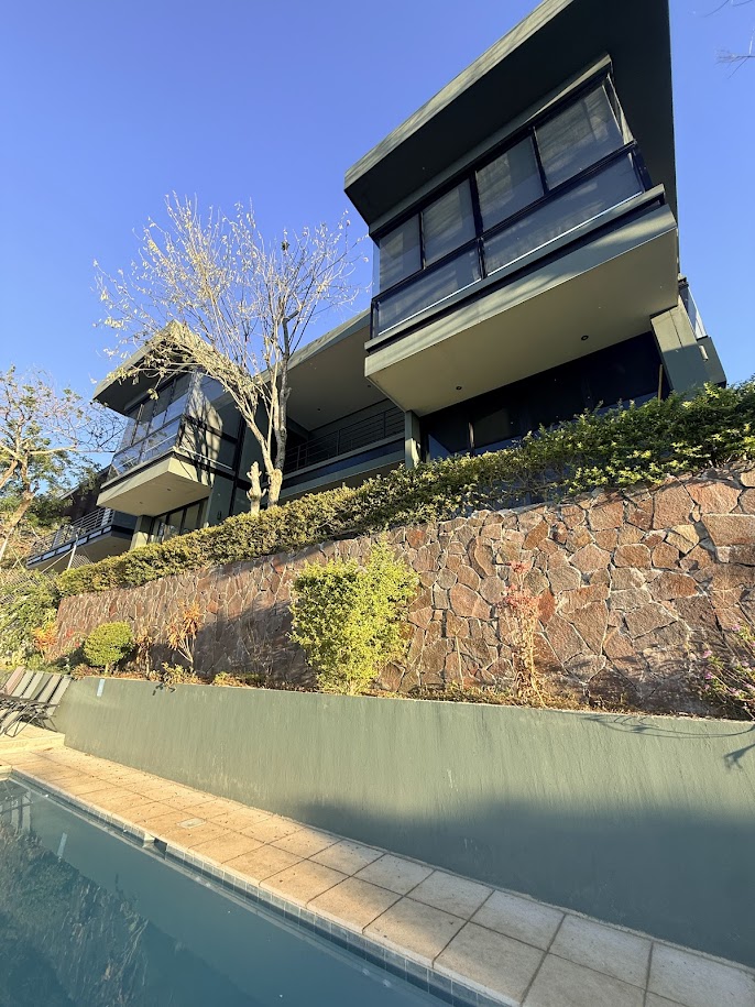 Modern multi-story house with large windows and balconies, situated on a hillside next to a swimming pool.