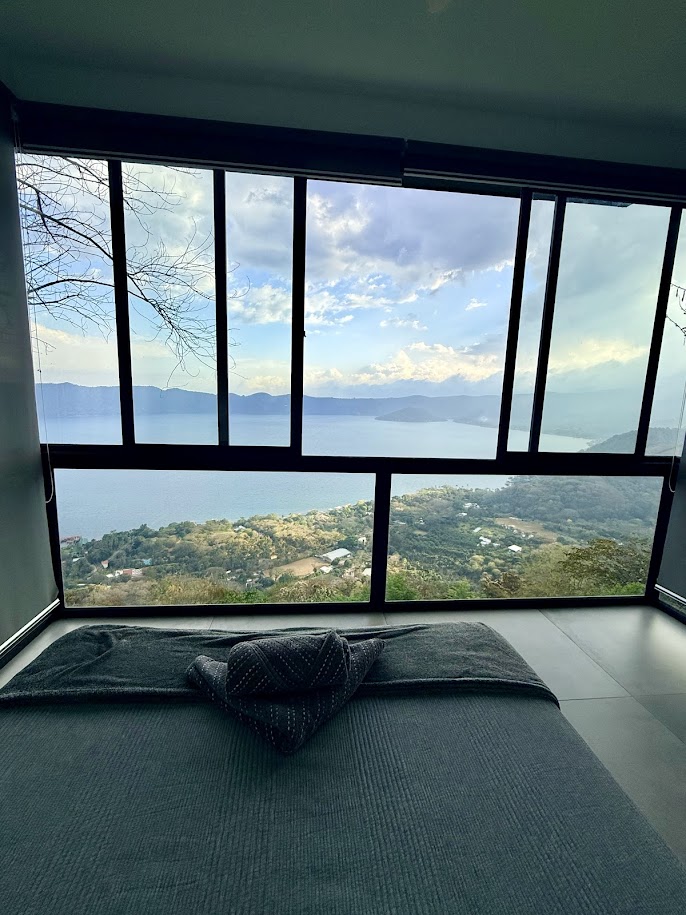 A cozy interior view from a room overlooking a stunning lake and mountains, with a neatly made bed in the foreground and large windows framing the scenery.