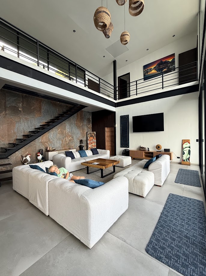 A spacious modern living room featuring a comfortable sectional sofa, wooden coffee table, and a large flat-screen TV. The room has high ceilings, wooden accents, and stylish hanging light fixtures. A child is seen relaxing on the sofa, with decorative art pieces and a staircase visible in the background.
