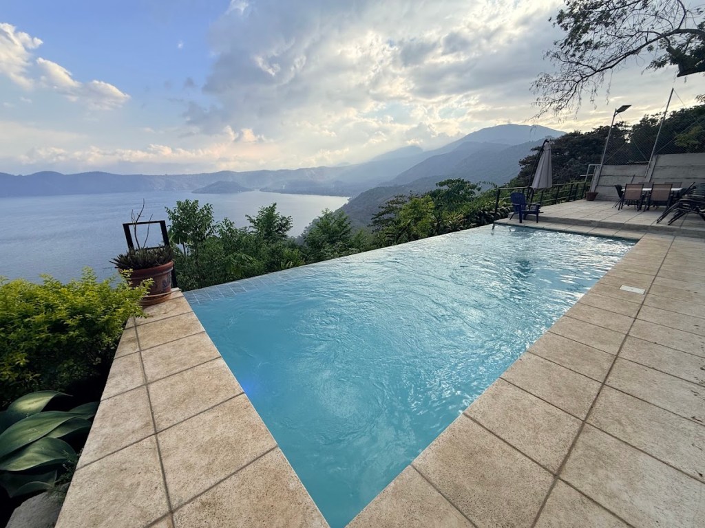 A serene infinity pool overlooking a lake and mountains, surrounded by lush greenery and a cloudy sky.