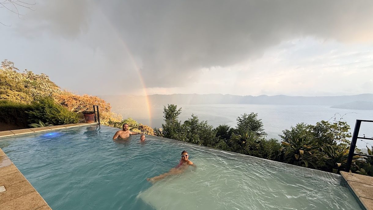 Two people relax in an infinity pool overlooking a scenic landscape with hills and a lake under a cloudy sky with a rainbow.
