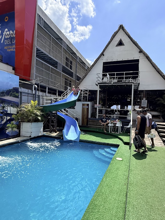 A vibrant pool area featuring a water slide, with a restaurant building in the background and people enjoying the space.