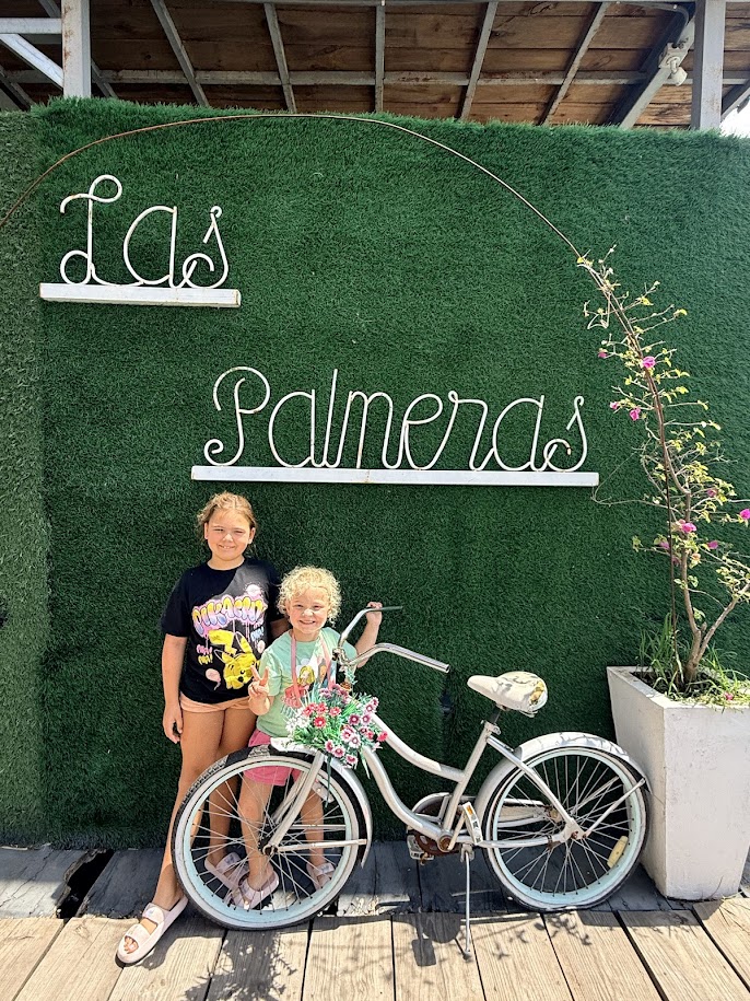 Two children standing next to a vintage bicycle decorated with flowers in front of a green backdrop with the words 'Las Palmeras' overhead.