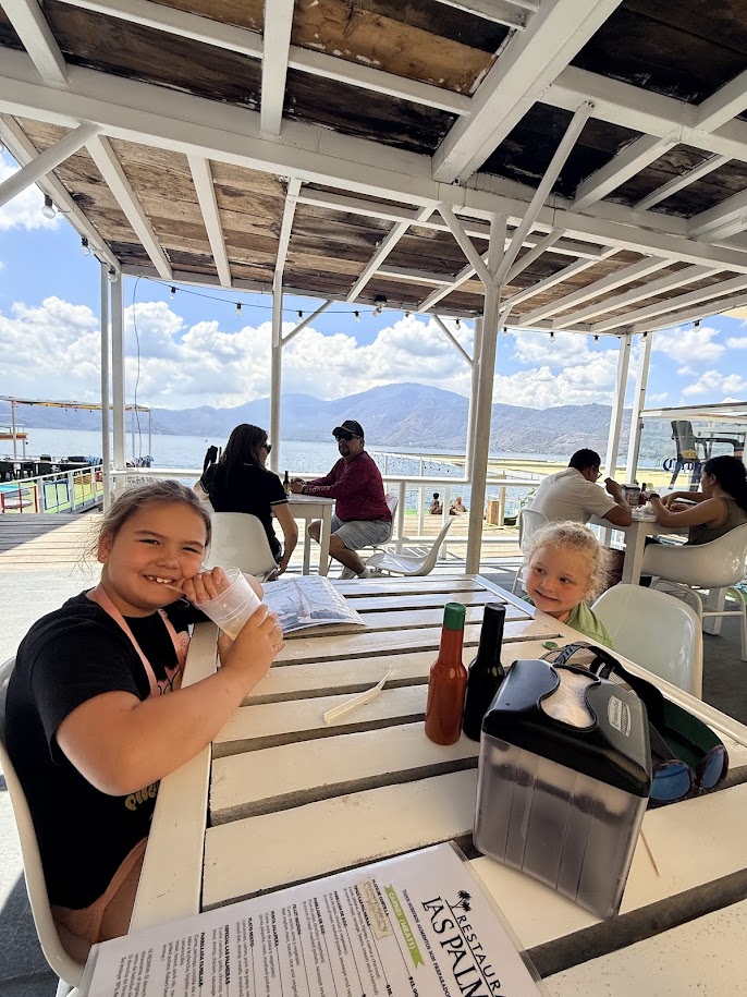 Two children sitting at a table in a restaurant, one smiling and holding a drink, with a view of a lake and mountains in the background.
