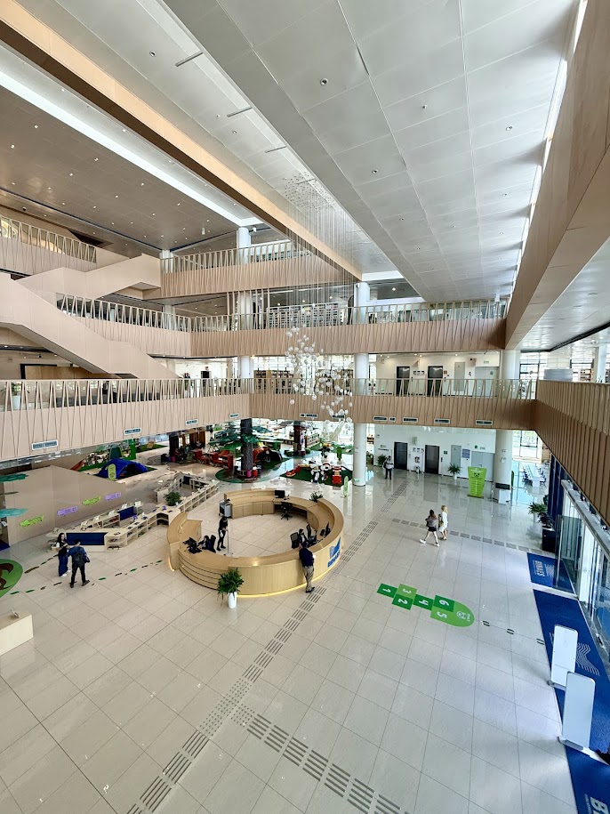 A spacious modern interior of a building with multiple levels, featuring wooden railings and a high ceiling. The central area includes a circular reception desk and various seating arrangements. People are seen walking and interacting in the space, which is well-lit and showcases greenery and a contemporary design.