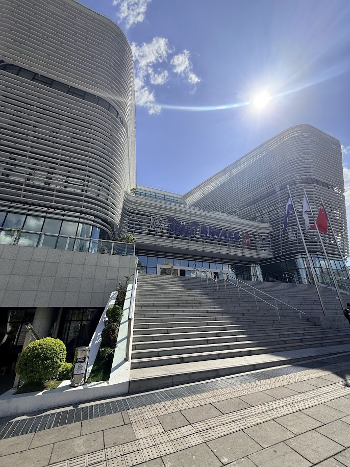 Modern architecture of a building with a glass facade, featuring wide stairs leading up to an entrance, illuminated by sunlight against a clear blue sky.