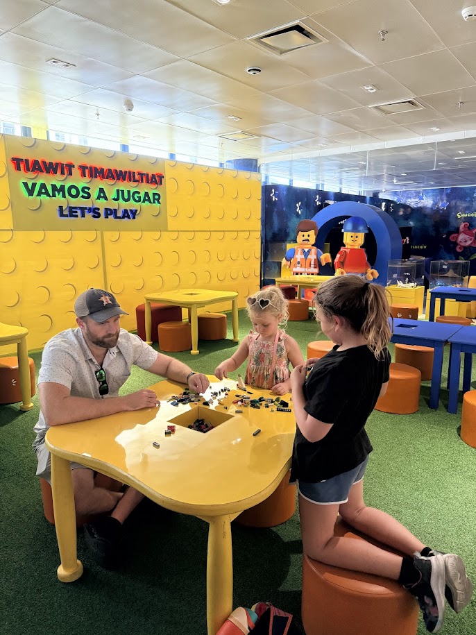 A man and two children playing with LEGO bricks at a bright yellow table in a colorful play area, with toys and playful decorations in the background.