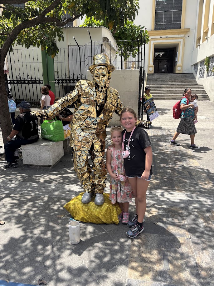 Two girls standing next to a golden statue performer in a busy outdoor setting.
