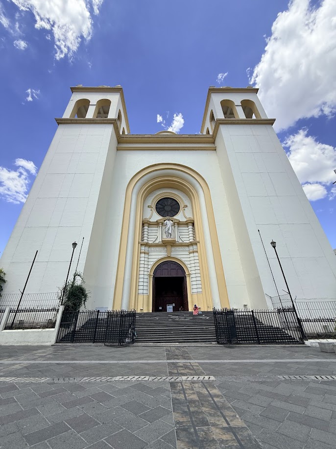 A front view of a large church featuring a grand entrance with steps, two bell towers, and a circular window, set against a blue sky with clouds.
