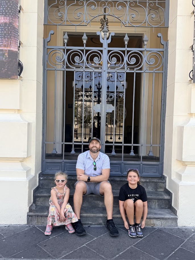 A man sitting on steps with two young girls in front of a decorative wrought iron gate.