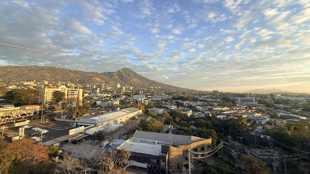 Panoramic view of a cityscape featuring mountains in the background under a cloudy sky, showcasing urban buildings and greenery.