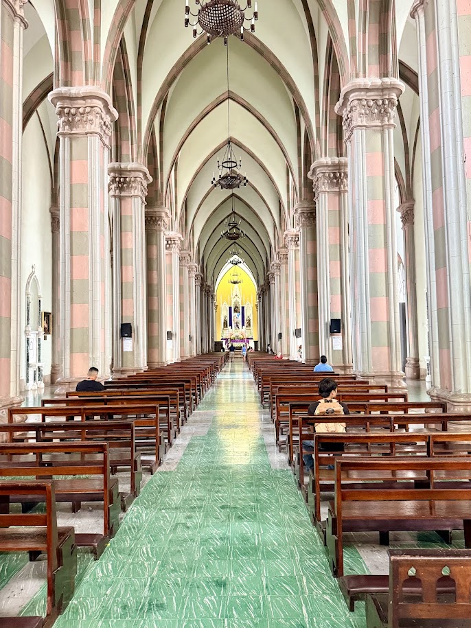 Interior view of a cathedral featuring tall arches, wooden benches, and a green aisle leading to an altar brightly lit at the end.