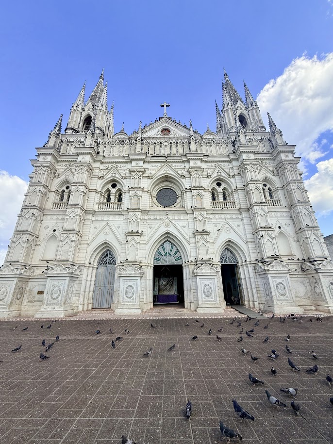 Front view of a large, ornate white cathedral with gothic architecture, featuring tall spires, arched doorways, and a round window, surrounded by pigeons on the ground under a blue sky.