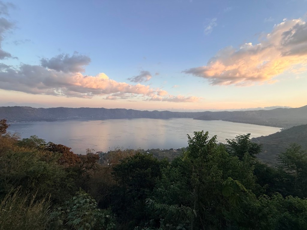 A scenic view of a lake surrounded by mountains at sunset, with colorful clouds reflecting on the water surface.