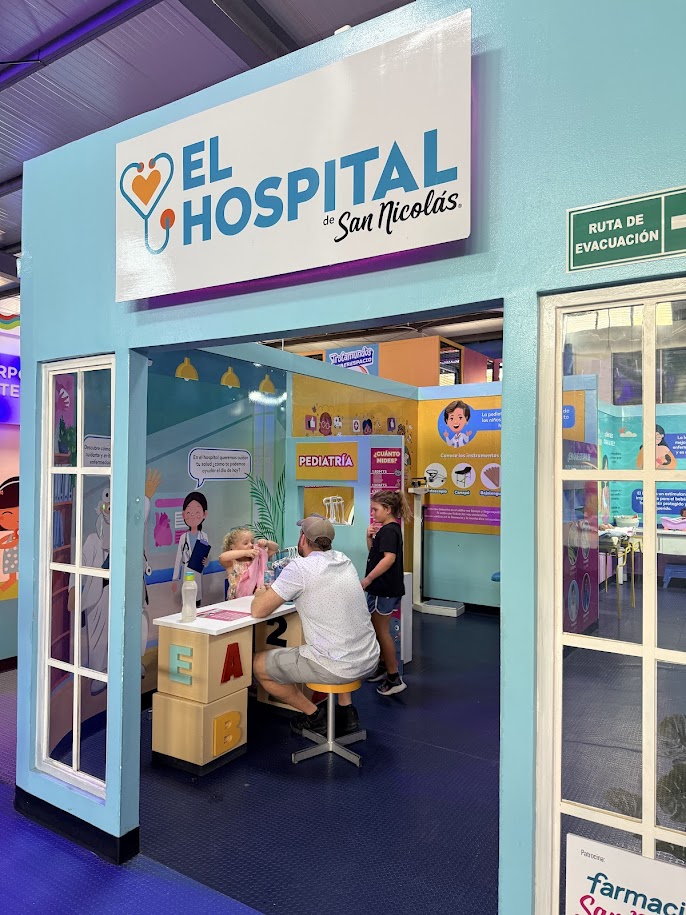 Entrance to 'El Hospital de San Nicolás' for children, featuring a sign and colorful displays related to pediatric care, with a man and two children engaging at a table.