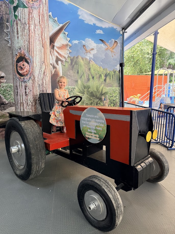 A young girl sits in a large, colorful toy tractor with a tree trunk backdrop and dinosaur-themed decorations in an outdoor play area.