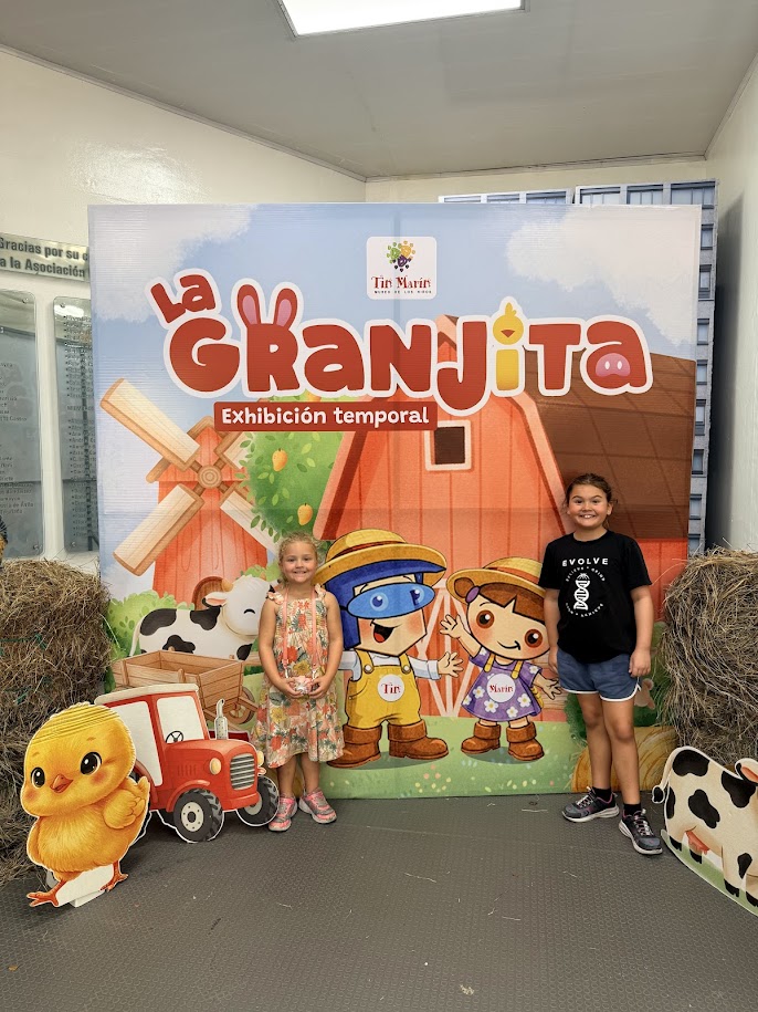 Two children posing in front of a colorful backdrop for 'La Granjita' temporary exhibition, featuring cartoon farm characters and props like a red tractor and hay bales.